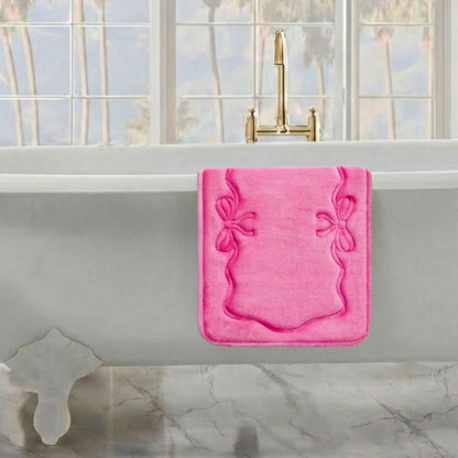 A Quilted Memory Foam Bath Mat (17" x 24") is draped over the edge of a white freestanding tub in a sunlit bathroom with marble floors, large windows, and a gold faucet. Palm trees can be seen just outside.