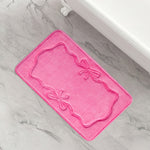 The Quilted Memory Foam Bath Mat, 20" x 32", in pink features embossed decorative borders and two bow designs, shown on a light gray marble floor beside a white bathtub.