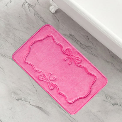 The Quilted Memory Foam Bath Mat, 20" x 32", in pink features embossed decorative borders and two bow designs, shown on a light gray marble floor beside a white bathtub.
