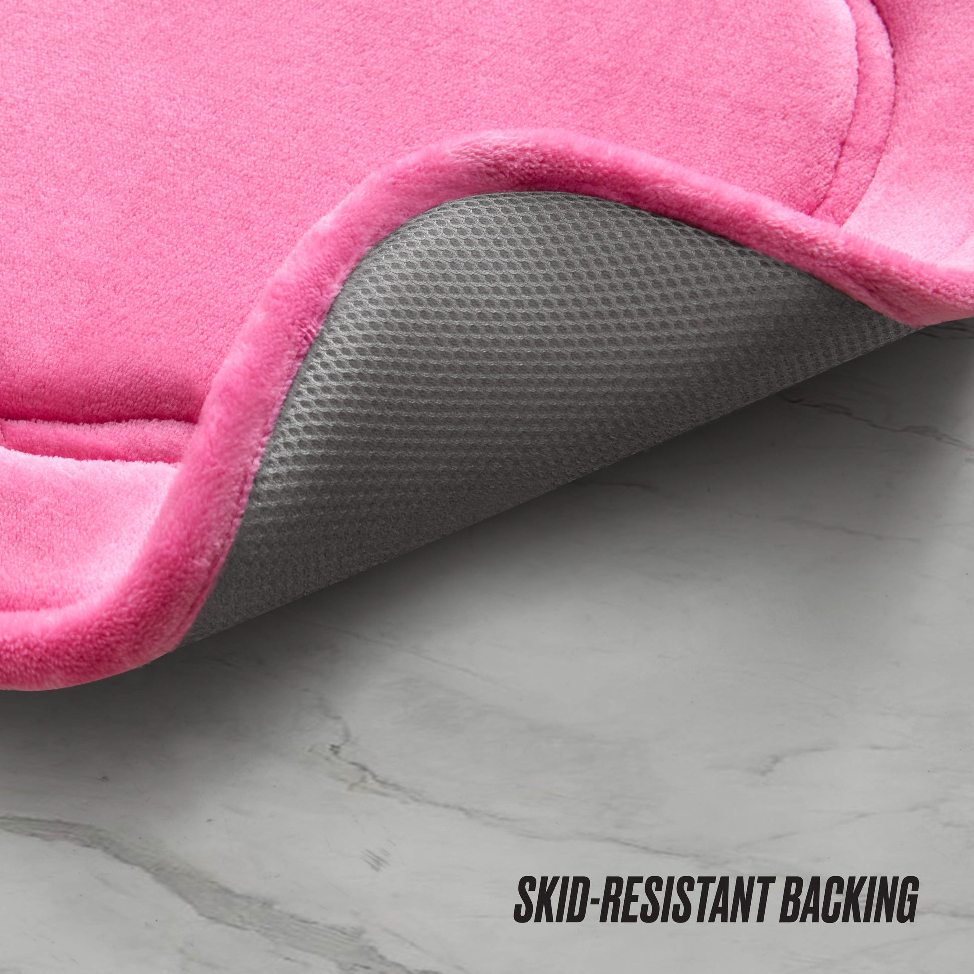 A close-up of the Quilted Memory Foam Bath Mat, 20" x 32", with a corner lifted to show its gray textured, skid-resistant backing. The pink quilted mat is on a light gray marble floor. Text: "SKID-RESISTANT BACKING.