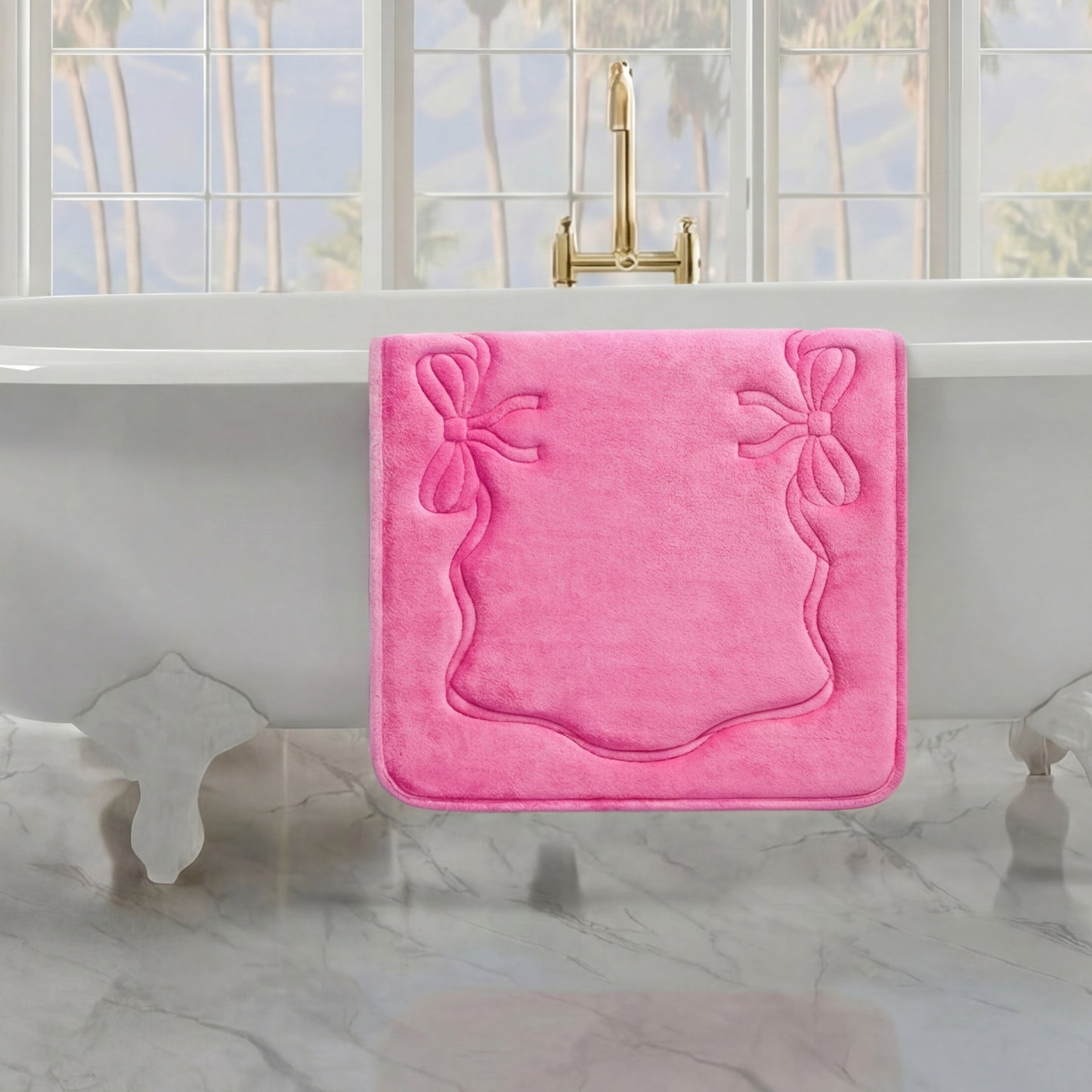 A pink Quilted Memory Foam Bath Mat, 20" x 32", hangs over the side of a white clawfoot tub in a bright bathroom with marble floors and large windows showcasing palm trees outside.