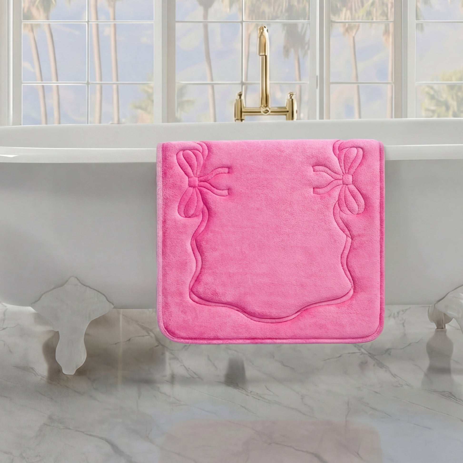 A pink Quilted Memory Foam Bath Mat, 20" x 32", hangs over the side of a white clawfoot tub in a bright bathroom with marble floors and large windows showcasing palm trees outside.