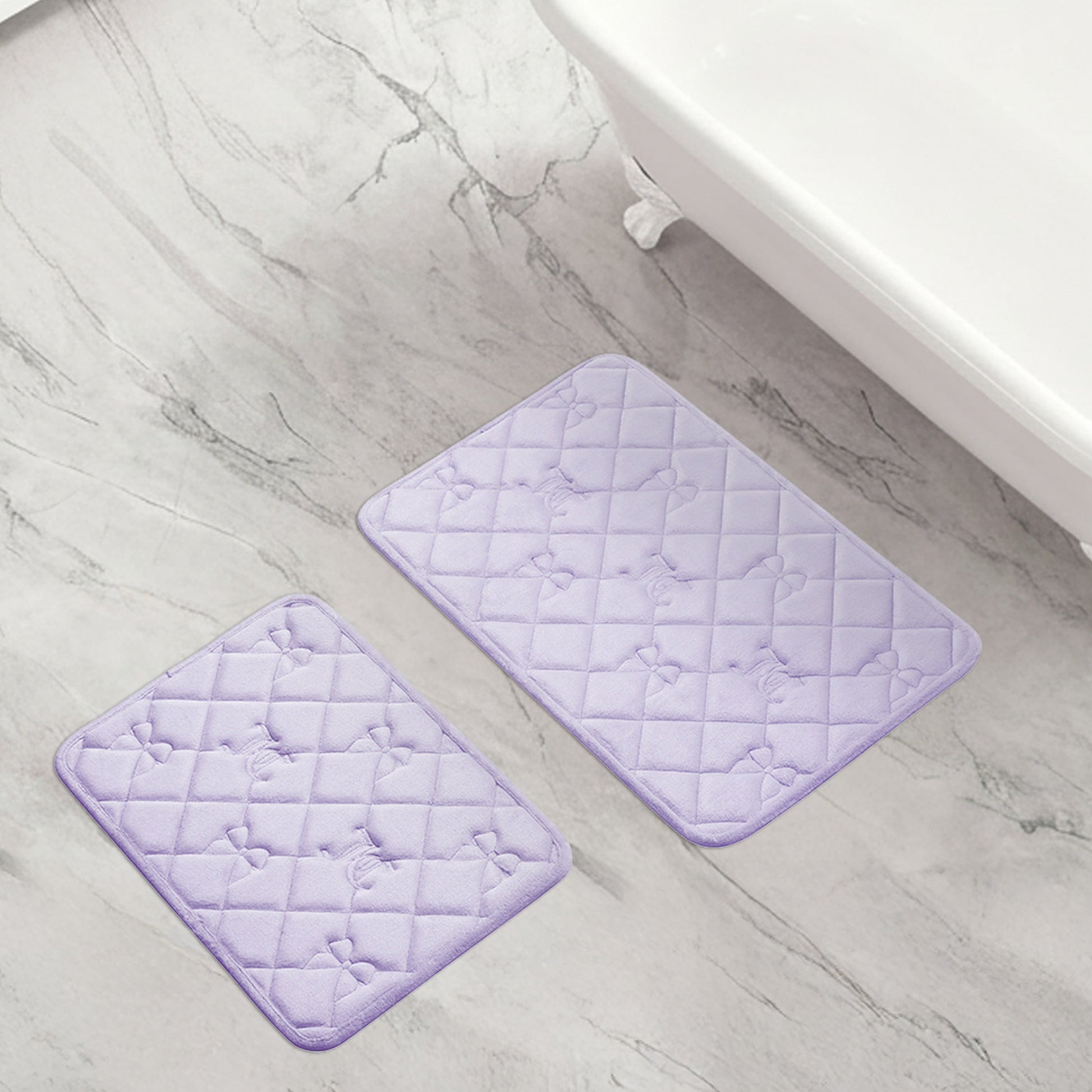 The Quilted Memory Foam Bath Mat 2-Piece Set in light purple, featuring a subtle bow pattern, is placed on a light gray marble-patterned bathroom floor next to a white bathtub.