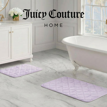 Elegant bathroom with white vanity, freestanding tub, and two light purple Quilted Memory Foam Bath Mats (2-Piece Set) on a marble floor. Large window in the background. Juicy Couture Home logo appears at the top.