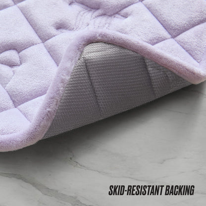 A close-up of the Quilted Memory Foam Bath Mat 2-Piece Set with lavender quilting, one corner lifted to show its textured gray skid-resistant backing on a light gray marble floor. "SKID-RESISTANT BACKING" appears in the corner.