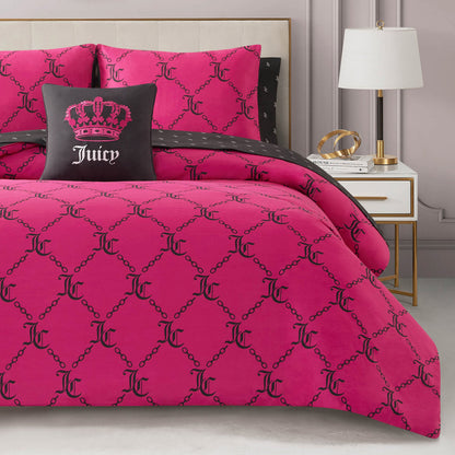 A bed dressed in the Link Printed Bed in a Bag Bedding Set, featuring black chain and JC patterns, is topped with a black pillow showing a pink crown and "Juicy." A white nightstand and lamp sit next to the bed, creating a luxurious atmosphere.