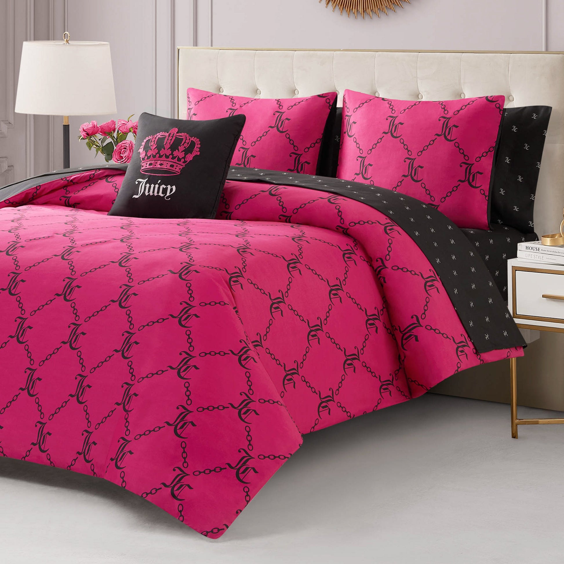 A stylish bedroom with the Link Printed Bed in a Bag Bedding Set—featuring a bright pink reversible comforter with black chain and logo patterns, matching pillows, a black crown accent pillow, and a modern nightstand with lamp and flowers.