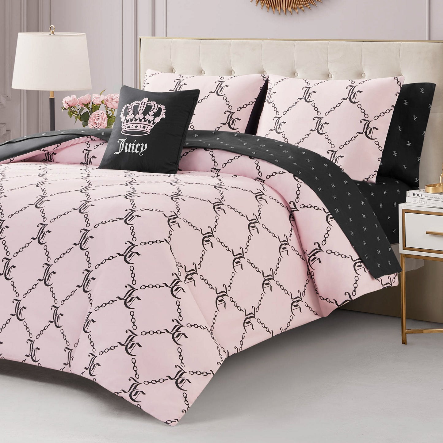 A neatly made bed with the Link Printed Bed in a Bag Bedding Set in pale pink, featuring a chain and JC pattern, black Juicy crown accent pillows, and a matching black sheet. A white lamp and pink flowers sit on the bedside table.