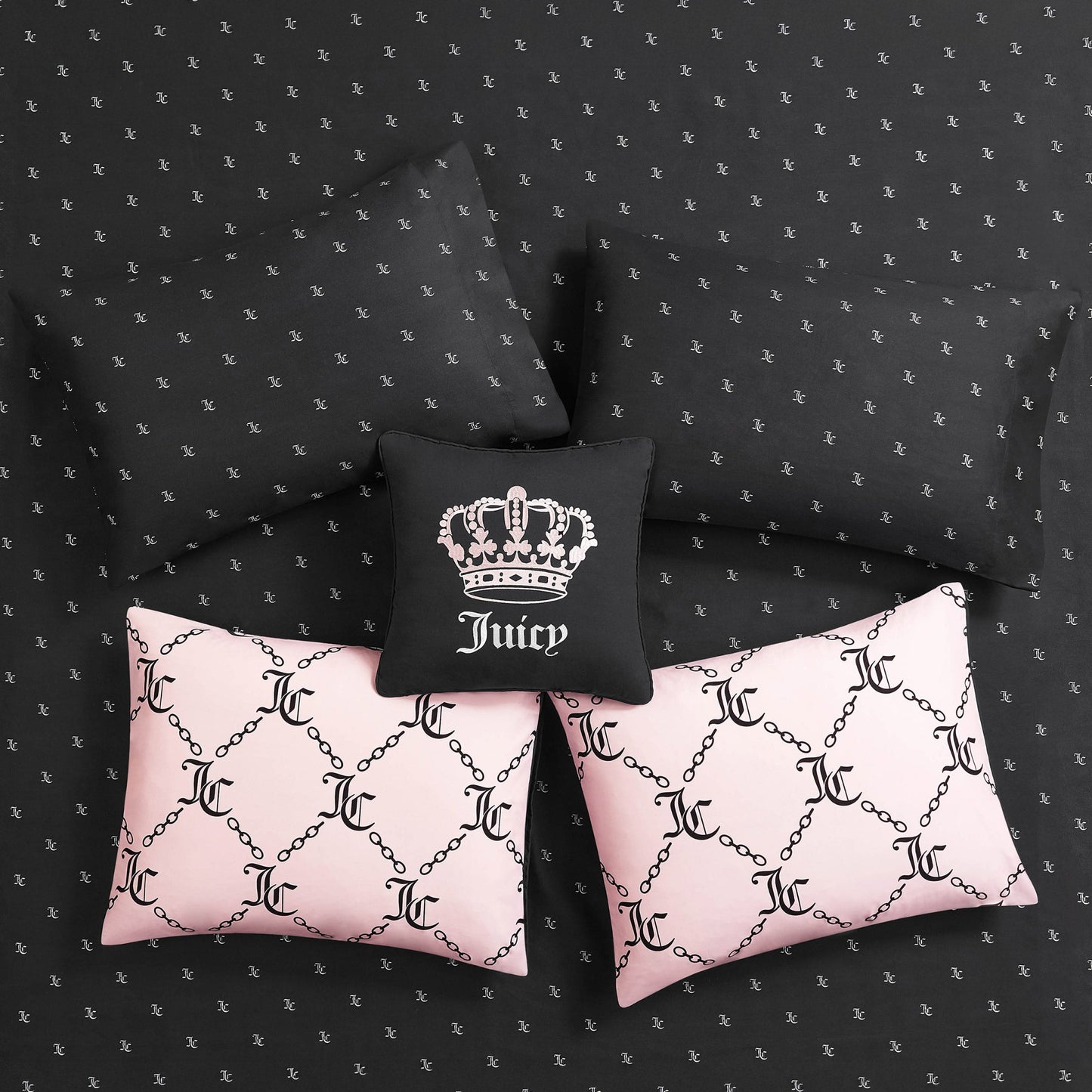Five pillows styled on the Link Printed Bed in a Bag Bedding Set: three black pillows (one with a white crown and "Juicy"), plus two light pink JC-logo pillows with chain patterns. The reversible bedding features a small repeating logo for luxe sleep.