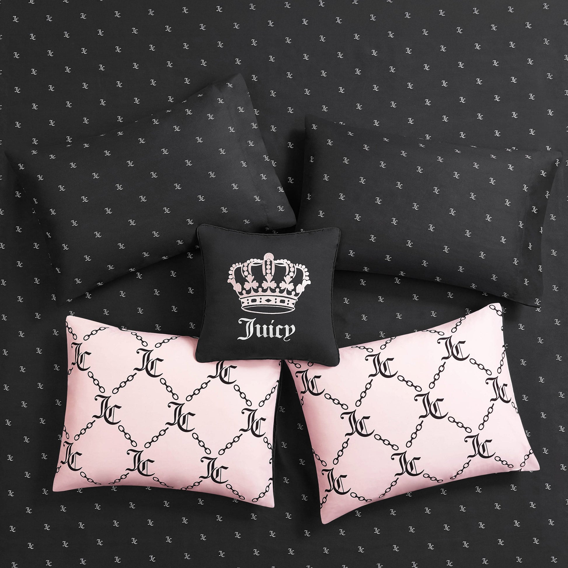 Five pillows styled on the Link Printed Bed in a Bag Bedding Set: three black pillows (one with a white crown and "Juicy"), plus two light pink JC-logo pillows with chain patterns. The reversible bedding features a small repeating logo for luxe sleep.