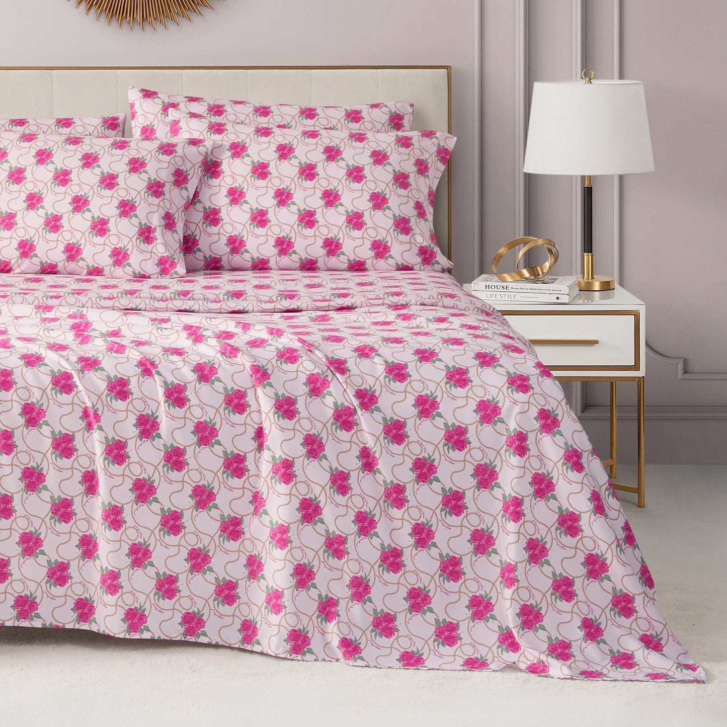A neatly made bed with a Microfiber Sheet Set With Extra Pillowcases and matching pillows features a white and pink floral bedspread. Next to the bed, a white nightstand with gold accents holds a modern lamp, books, and décor against light gray walls.