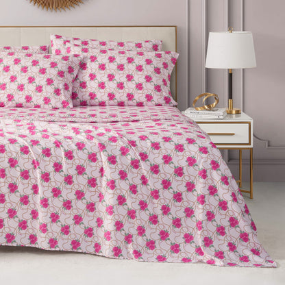 A neatly made bed with a Microfiber Sheet Set With Extra Pillowcases and matching pillows features a white and pink floral bedspread. Next to the bed, a white nightstand with gold accents holds a modern lamp, books, and décor against light gray walls.
