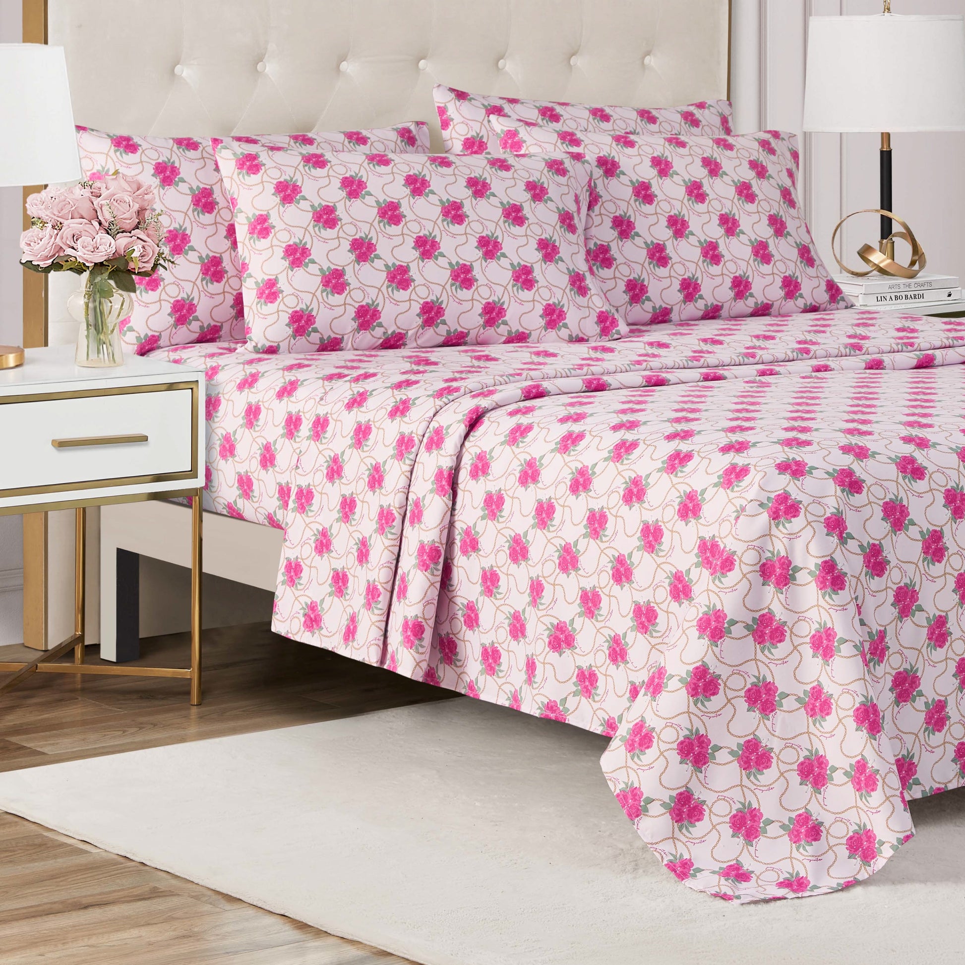 A neatly made bed with a pink and white floral-patterned Microfiber Sheet Set With Extra Pillowcases in a modern bedroom featuring a white nightstand, gold lamp, pink flowers in a vase, neutral rug, and wooden floor.