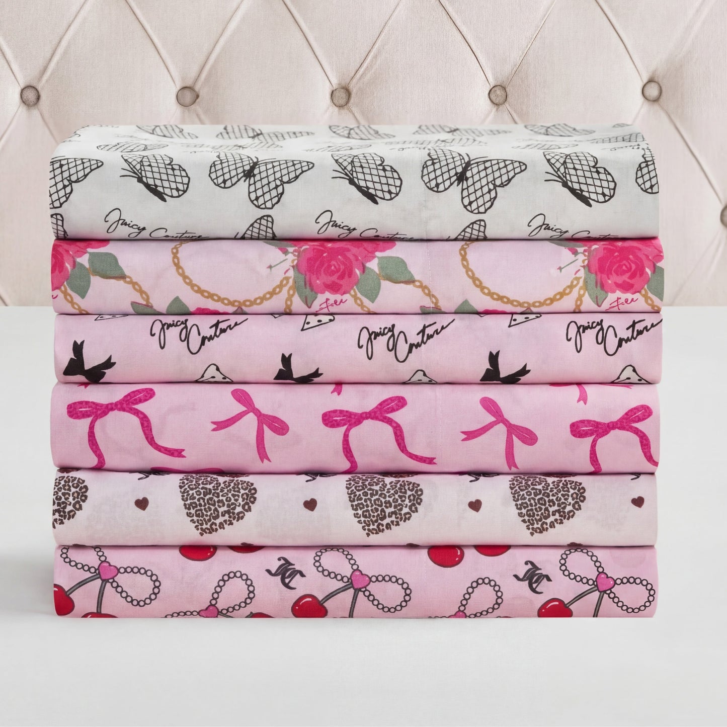 A stack of five Microfiber Sheet Set With Extra Pillowcases in shades of white and pink with heart, bow, rose, cherry, and bird prints is neatly arranged on a white surface—ideal for luxury bedding lovers.