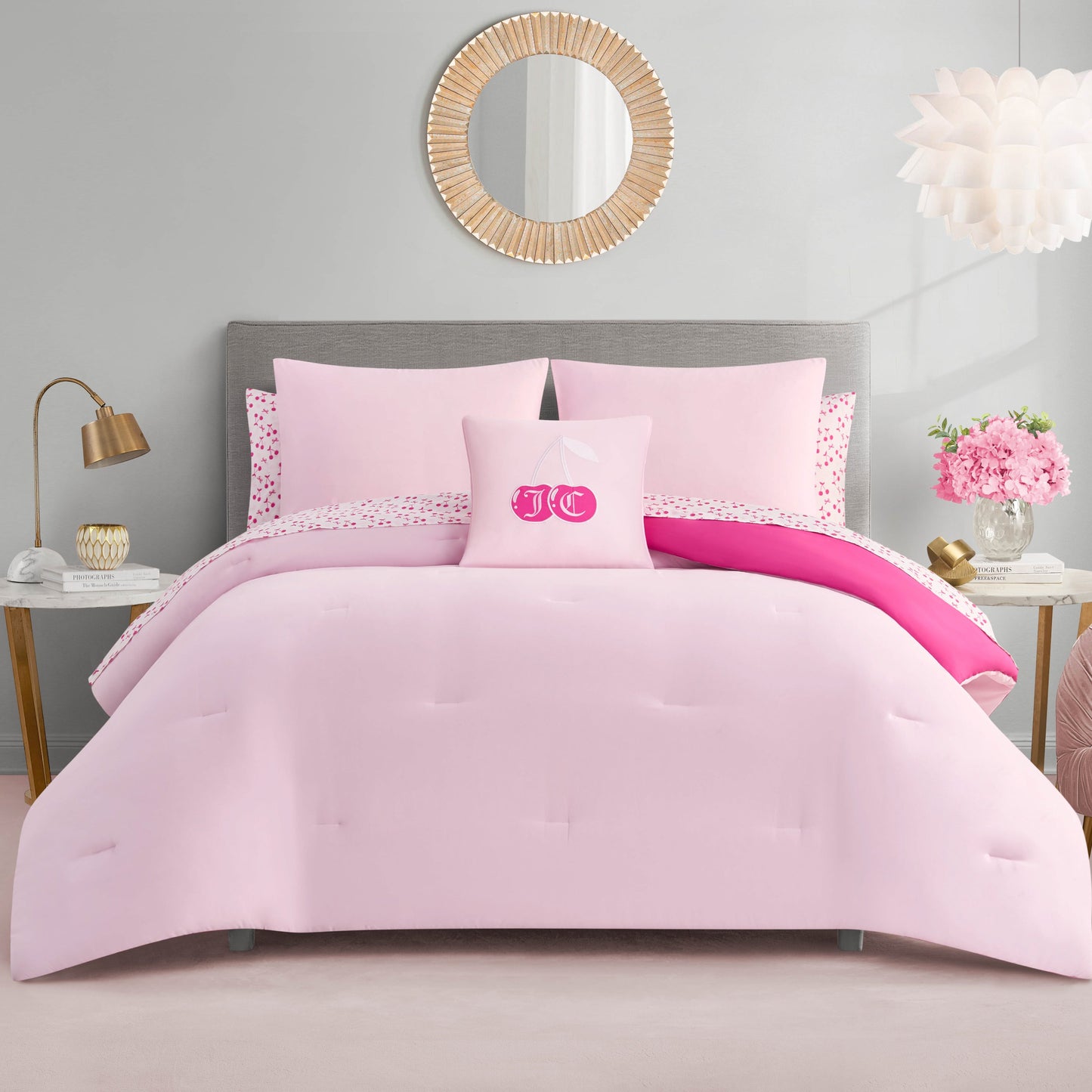 A neatly made bed with the Cherry Reversible Bed in a Bag Bedding Set and decorative pillows, including a monogrammed one in the center. The room features gold accents, a round mirror, flowers, and soft neutral walls.