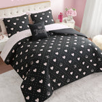 A neatly made bed features the Diamond Hearts Black Comforter Bed in a Bag Bedding Set, with a black comforter and pillowcases adorned with white hearts, crowns, and polka dots. A decorative pillow says "Juicy." Pink walls and floral decor add glam style.