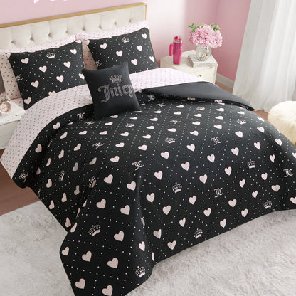 A neatly made bed features the Diamond Hearts Black Comforter Bed in a Bag Bedding Set, with a black comforter and pillowcases adorned with white hearts, crowns, and polka dots. A decorative pillow says "Juicy." Pink walls and floral decor add glam style.