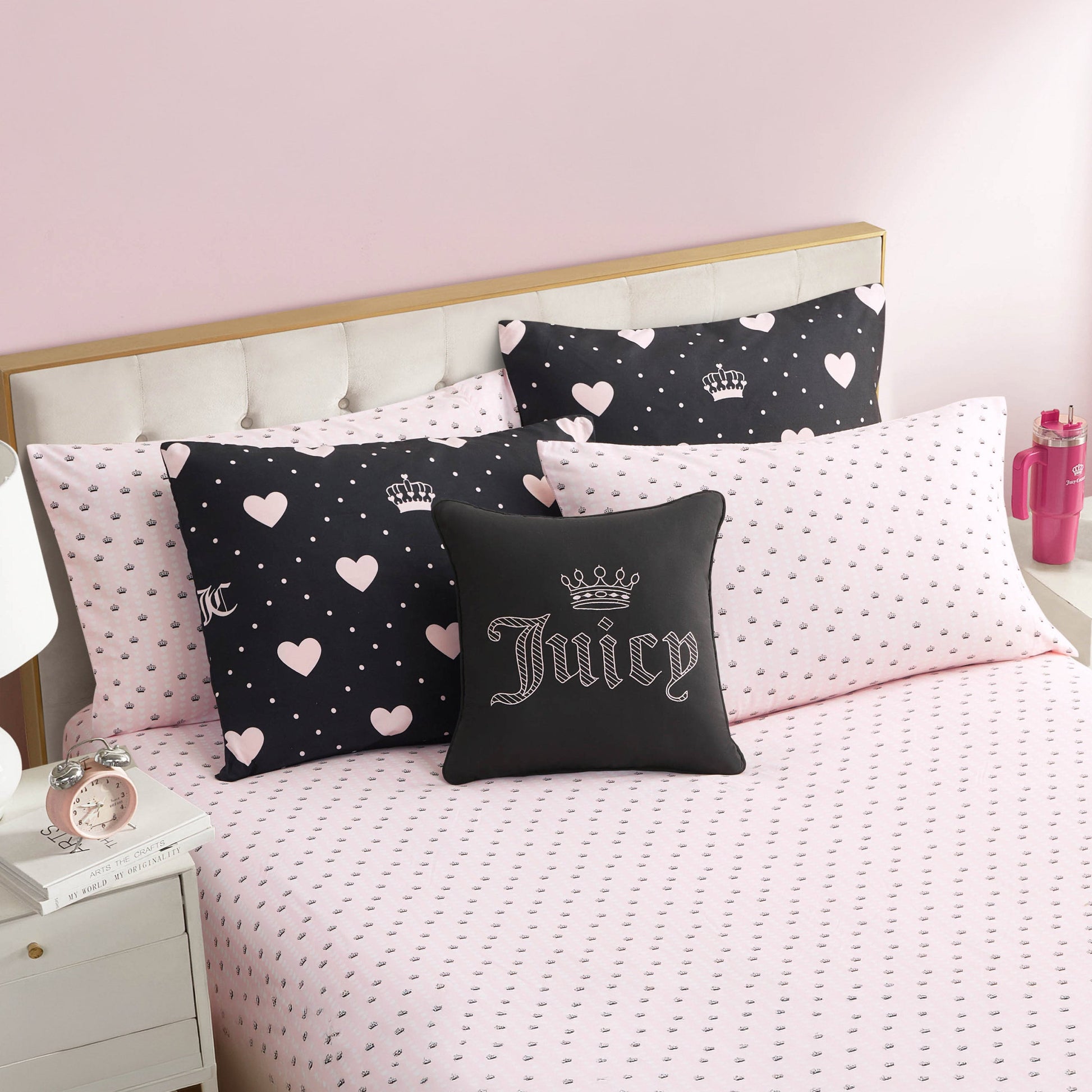 A neatly made bed with the Diamond Hearts Black Comforter Bed in a Bag Set, featuring pink heart-patterned sheets and glam black and pink pillows. A white tufted headboard, pink wall, and nightstand complete the stylish look.