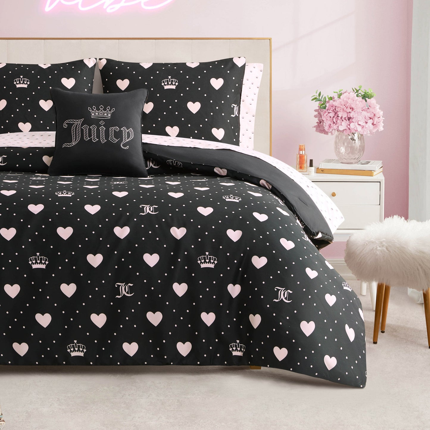 A neatly made bed features the Diamond Hearts Black Comforter Bed in a Bag Set, adorned with pink hearts, crowns, and JC logos. A black "Juicy" pillow with a crown sits center stage, popping against pink walls near flowers and a plush stool.