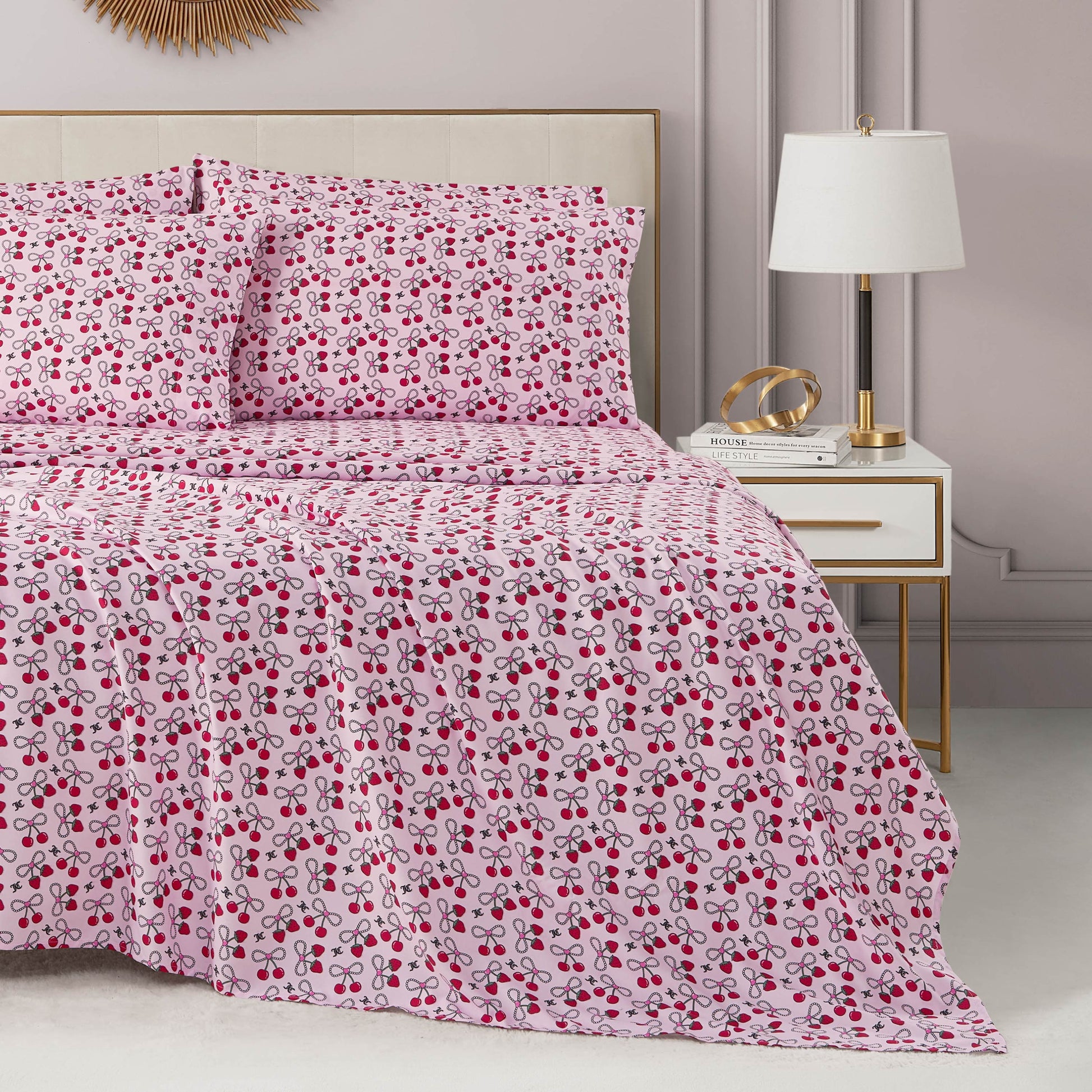 A neatly made bed with a pink, red, and purple cherry-patterned Microfiber Sheet Set With Extra Pillowcases sits beside a white nightstand with gold accents, a modern lamp, decorative items, and a soft pink wall in the background.