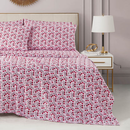 A neatly made bed with a pink, red, and purple cherry-patterned Microfiber Sheet Set With Extra Pillowcases sits beside a white nightstand with gold accents, a modern lamp, decorative items, and a soft pink wall in the background.