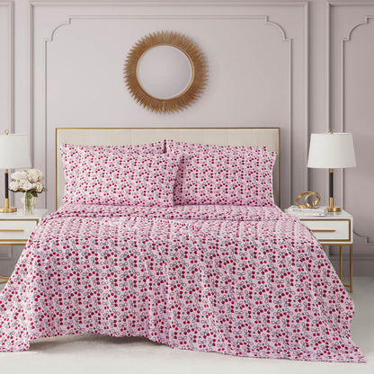 A bed with a Microfiber Sheet Set With Extra Pillowcases in pink and red floral patterns sits between two white nightstands with lamps against a light pink paneled wall, with a round gold sunburst mirror above the bed.