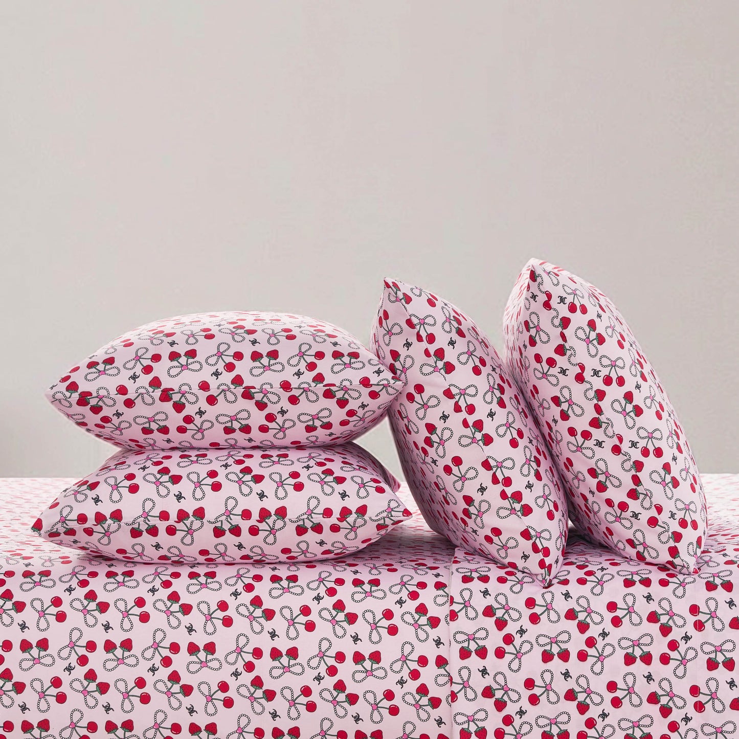 Four pillows and a bed are covered with white fabric featuring a red cherry and gray stem print, set against a light grey wall—an ideal look for the Microfiber Sheet Set With Extra Pillowcases or any luxury bedding collection.