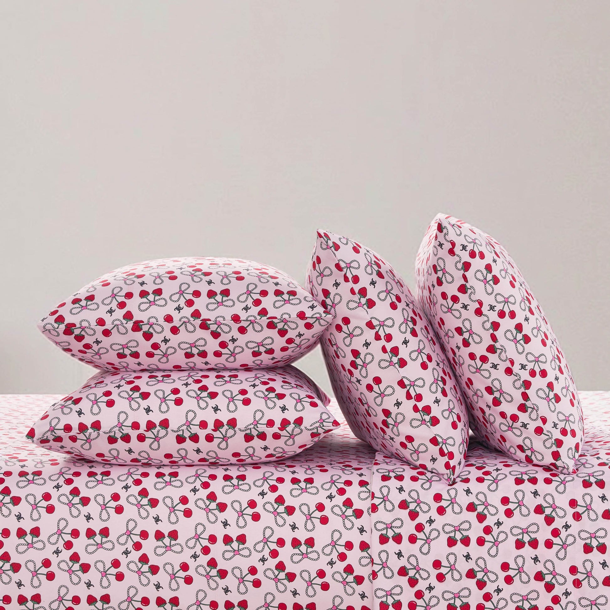 Four pillows and a bed are covered with white fabric featuring a red cherry and gray stem print, set against a light grey wall—an ideal look for the Microfiber Sheet Set With Extra Pillowcases or any luxury bedding collection.