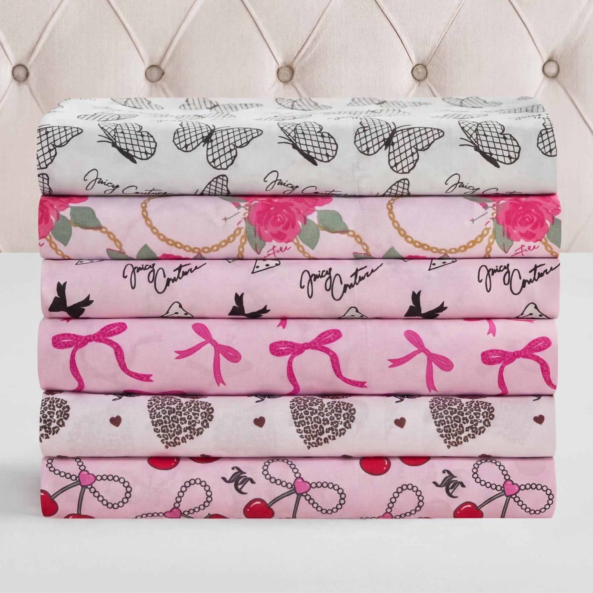 Six folded patterned sheets in pink, white, and gray—featuring hearts, bows, flowers, chains, birds, and cherries—this Microfiber Sheet Set With Extra Pillowcases adds playful charm to your luxury bedding collection. Neatly stacked before a tufted headboard.