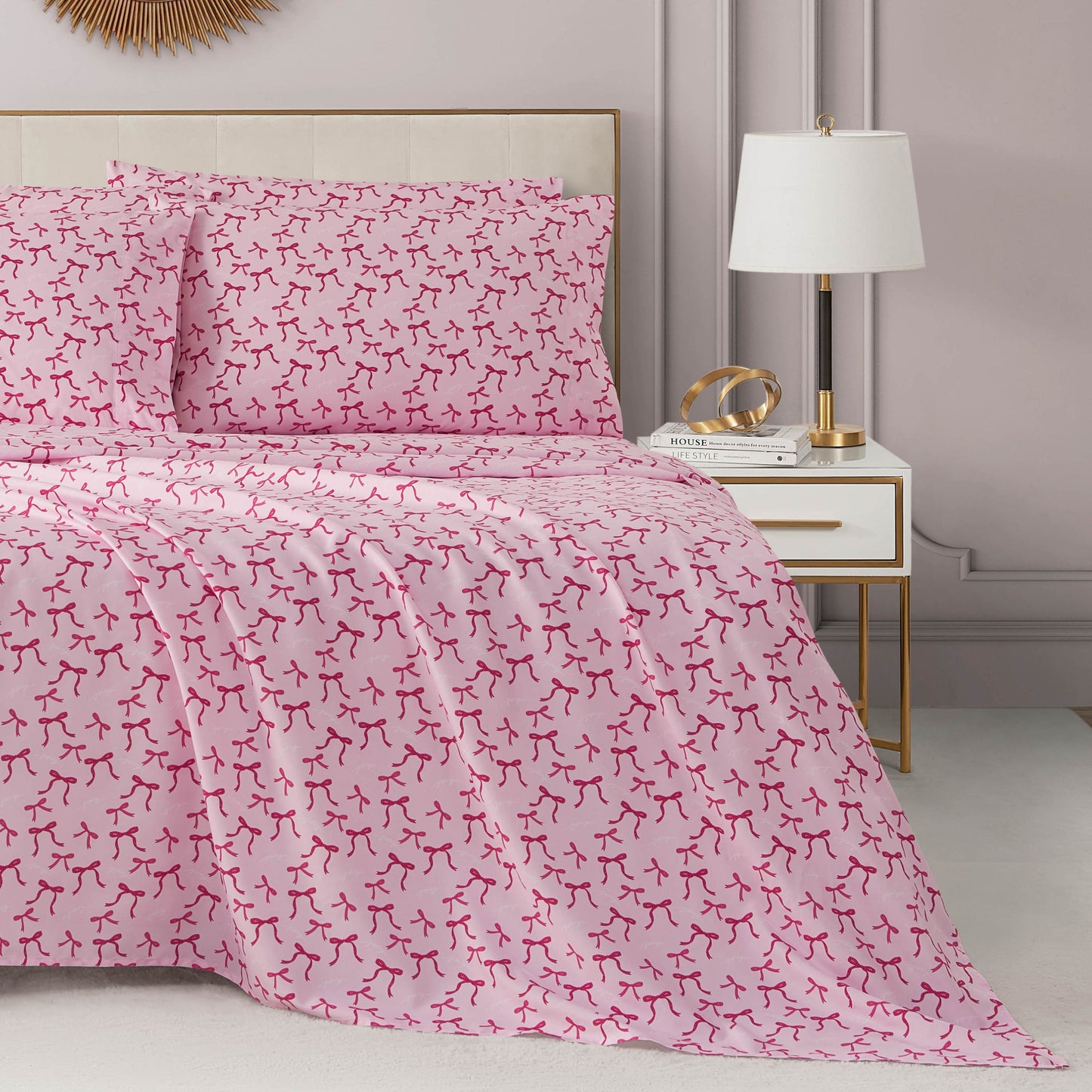 A neatly made bed with a Microfiber Sheet Set With Extra Pillowcases in pink and red abstract patterns sits next to a white and gold nightstand topped with a lamp and decor in a modern, softly lit bedroom.