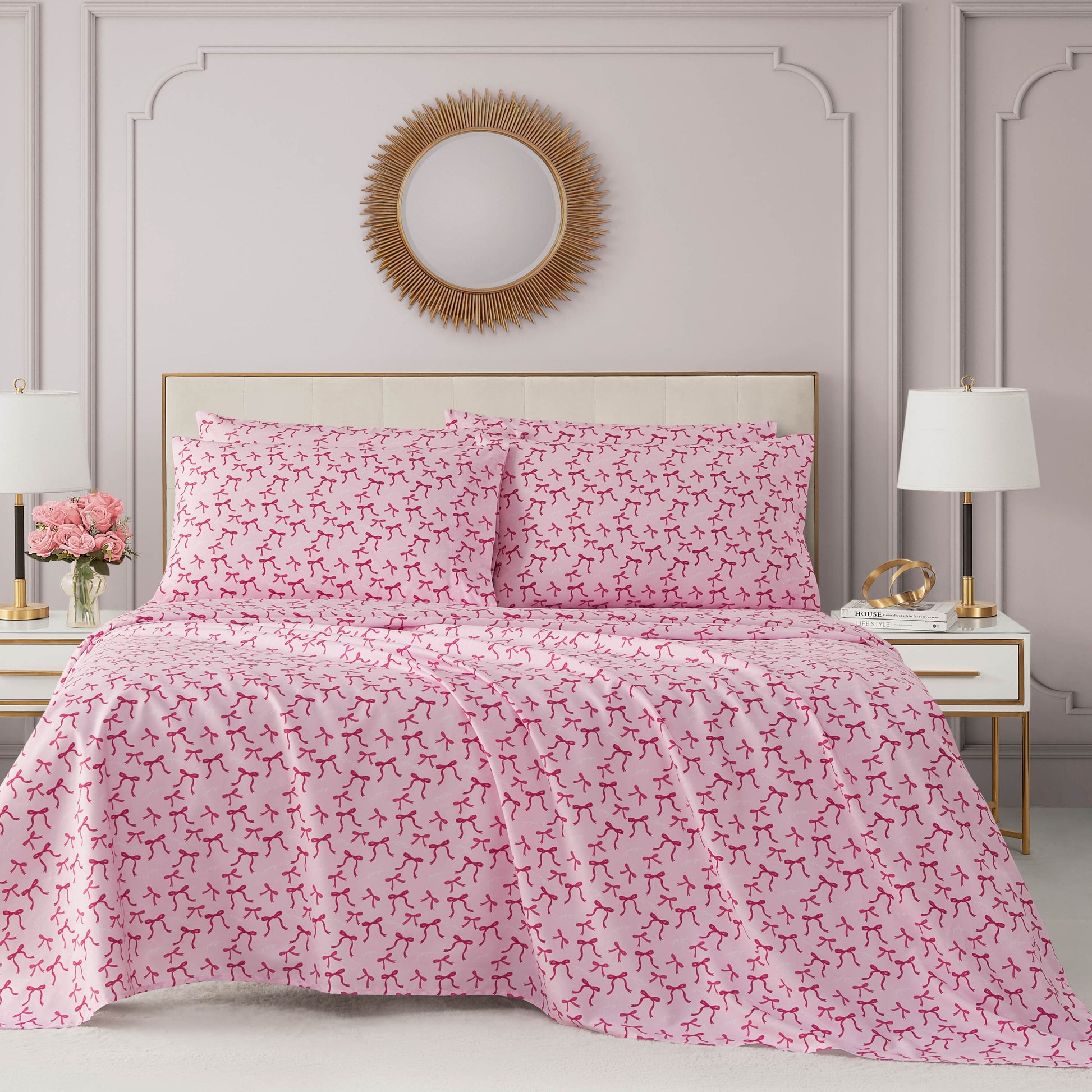 A neatly made bed with a pink and white comforter and pillows, flanked by two white nightstands with lamps and decor. Above the Microfiber Sheet Set With Extra Pillowcases hangs a round mirror with a gold sunburst frame.