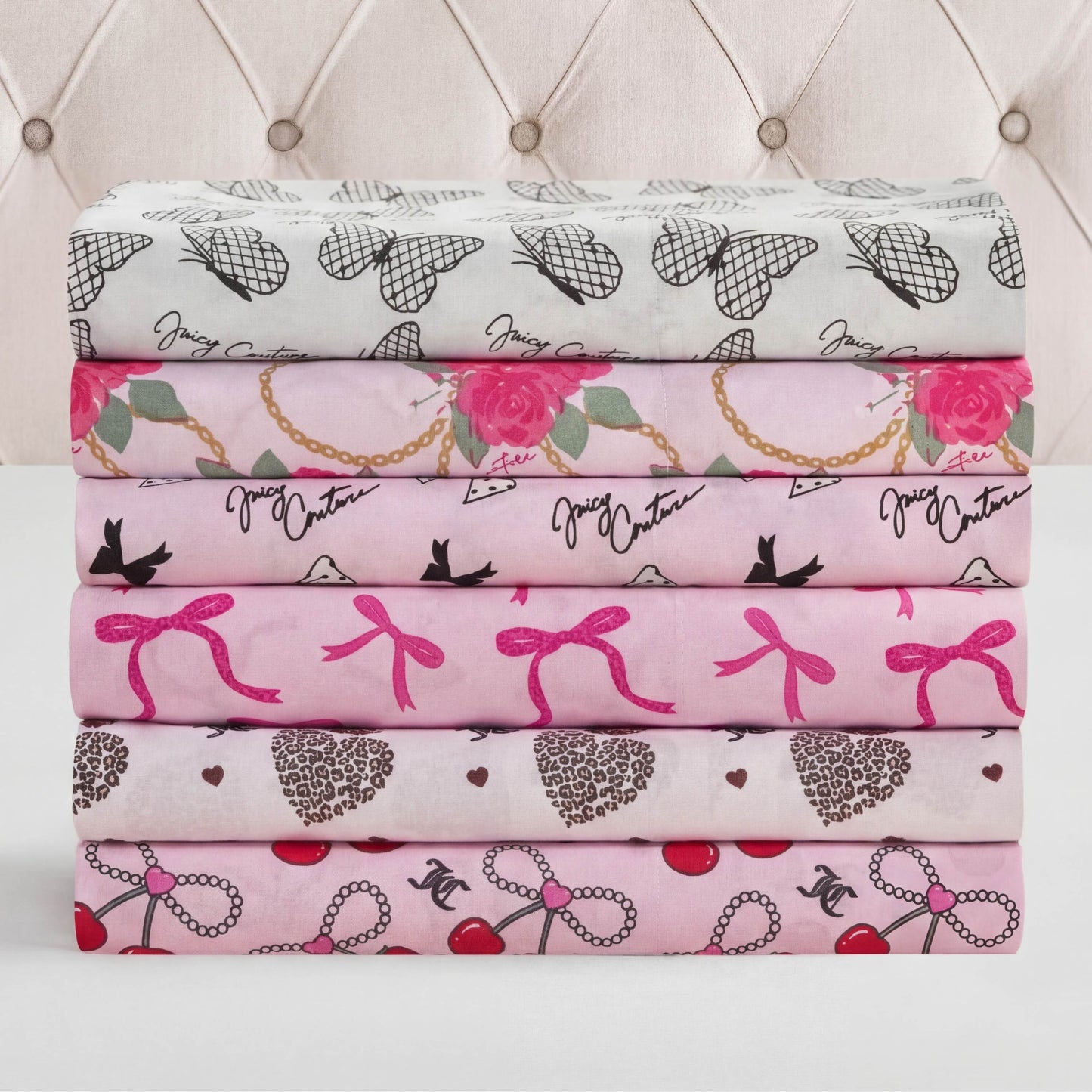 Six folded Microfiber Sheet Set With Extra Pillowcases, featuring pink and white prints like hearts, bows, cherries, and script, are stacked before a tufted beige headboard—bringing a touch of luxury bedding to any room.