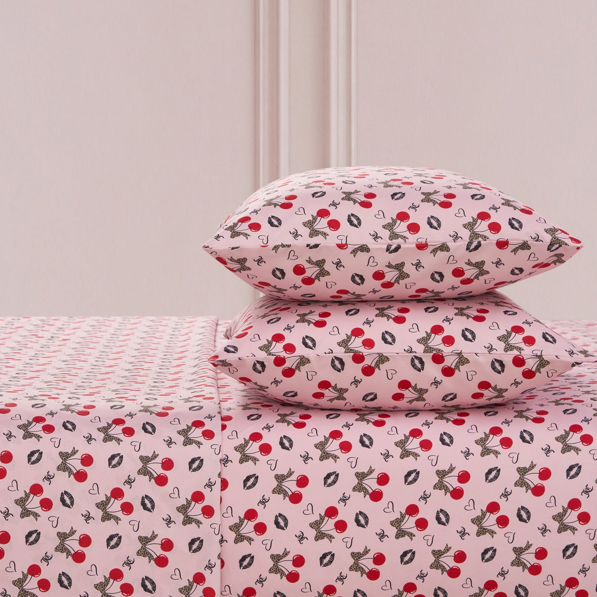 The Printed Microfiber Sheet Set includes two pillows and a sheet, all featuring red cherries, black and white bunnies, hearts, and white flowers on a soft pink background—a playful yet luxurious bedding choice.