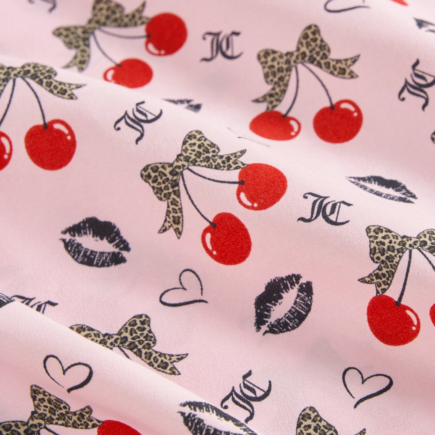 The Printed Microfiber Sheet Set features pink fabric with red cherries, leopard print bows, black lips, hearts, and JC letters for a playful look. Enjoy soft folds and stylish luxury bedding in any room.