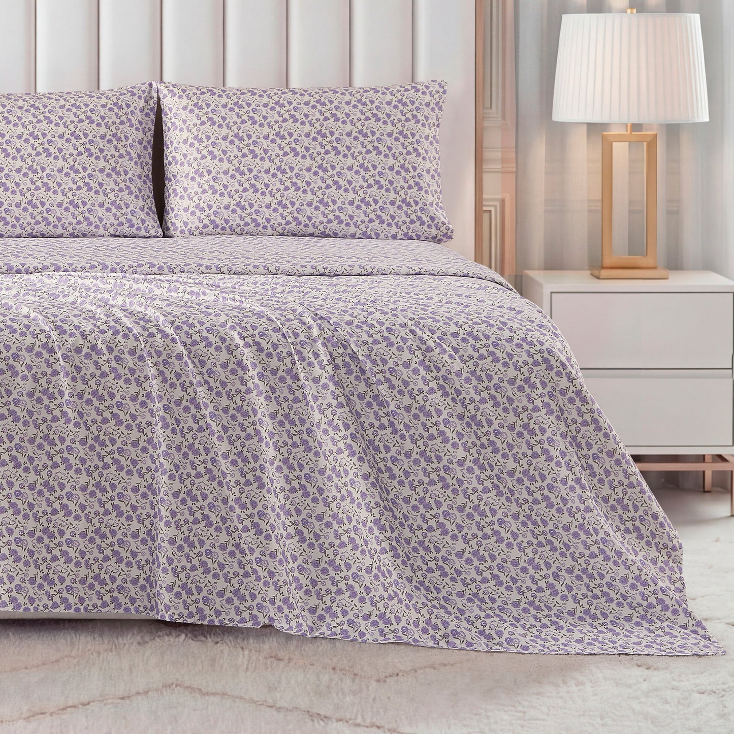 A bed dressed in the Printed Microfiber Sheet Set, featuring a purple and white floral pattern with matching pillowcases, sits beside a white nightstand and gold-base lamp in a warmly lit bedroom.