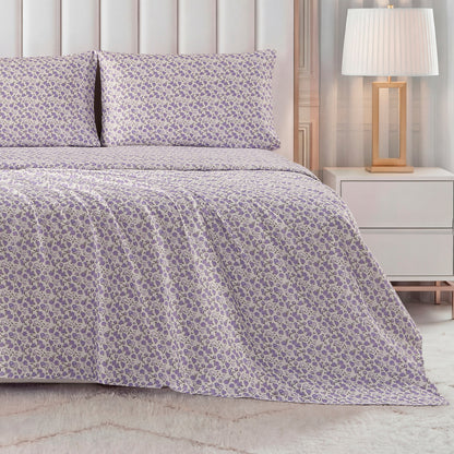 A bed dressed in the Printed Microfiber Sheet Set, featuring a purple and white floral pattern with matching pillowcases, sits beside a white nightstand and gold-base lamp in a warmly lit bedroom.