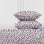 Two pillows and a Printed Microfiber Sheet Set, all with a small purple floral pattern on a light background, are stacked together in front of a minimal white wall.