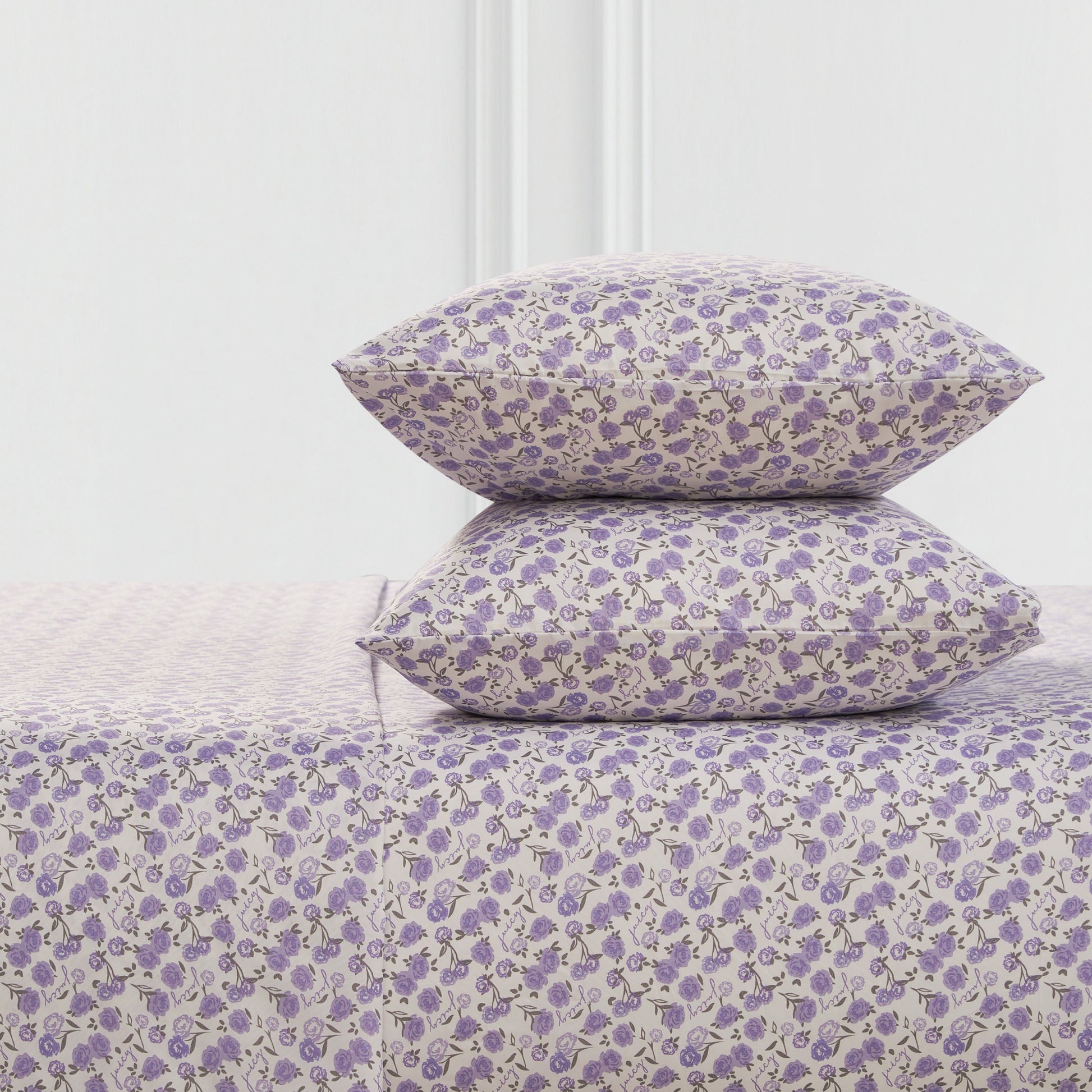 Two pillows and a Printed Microfiber Sheet Set, all with a small purple floral pattern on a light background, are stacked together in front of a minimal white wall.