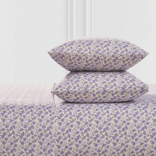 Two pillows and a Printed Microfiber Sheet Set, all with a small purple floral pattern on a light background, are stacked together in front of a minimal white wall.
