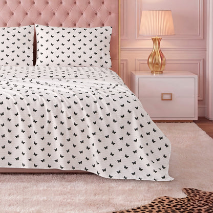 A bed dressed in the Printed Microfiber Sheet Set showcases a black butterfly pattern, with a tufted pink headboard, gold lamp on a white nightstand, light pink wall, and part of a leopard print rug at the foot of the bed.