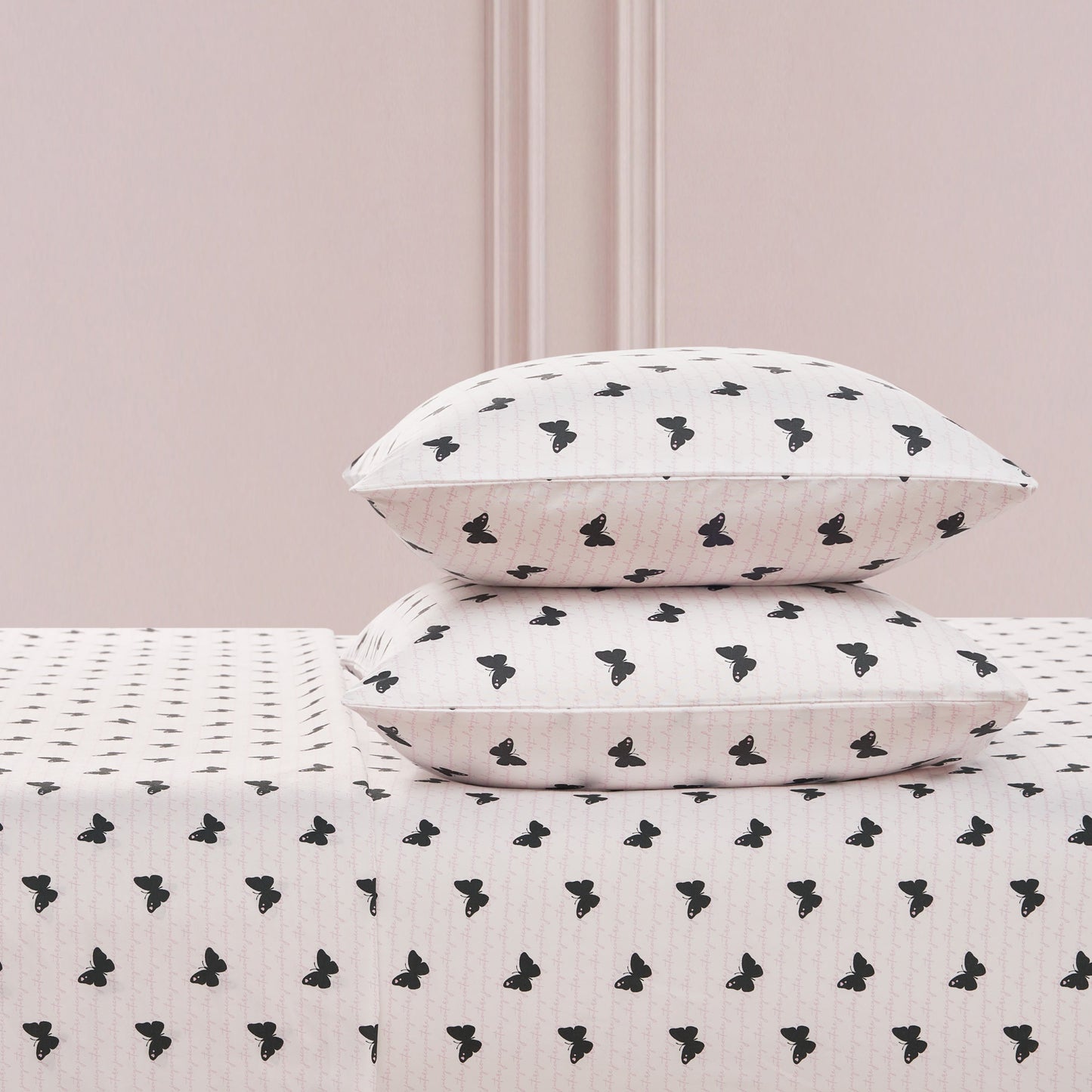 Two white pillows with a black butterfly print are stacked on the Printed Microfiber Sheet Set, set against a light pink wall with decorative molding.