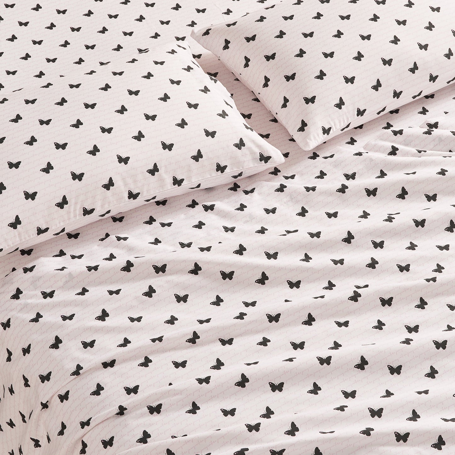 A close-up of a bed with two pillows and a blanket, all covered in the Printed Microfiber Sheet Set featuring white fabric with a repeating pattern of small black butterflies.