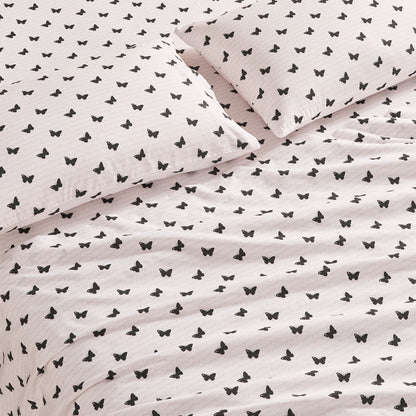 A close-up of a bed with two pillows and a blanket, all covered in the Printed Microfiber Sheet Set featuring white fabric with a repeating pattern of small black butterflies.