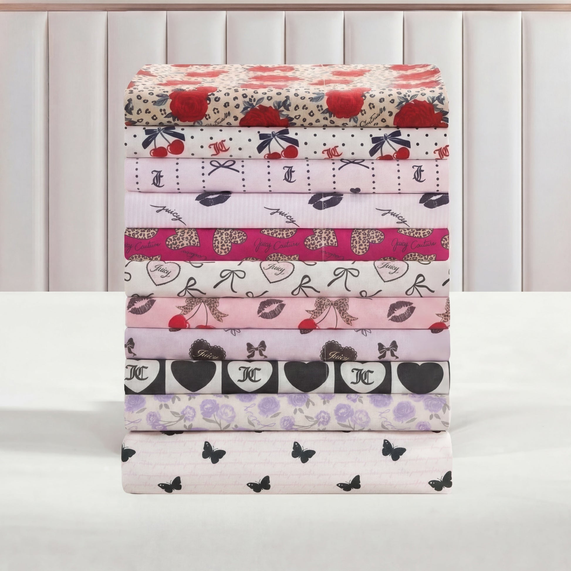 A stack of neatly folded Printed Microfiber Sheet Sets with heart, bow, and butterfly prints is arranged on a white surface against a padded backdrop.