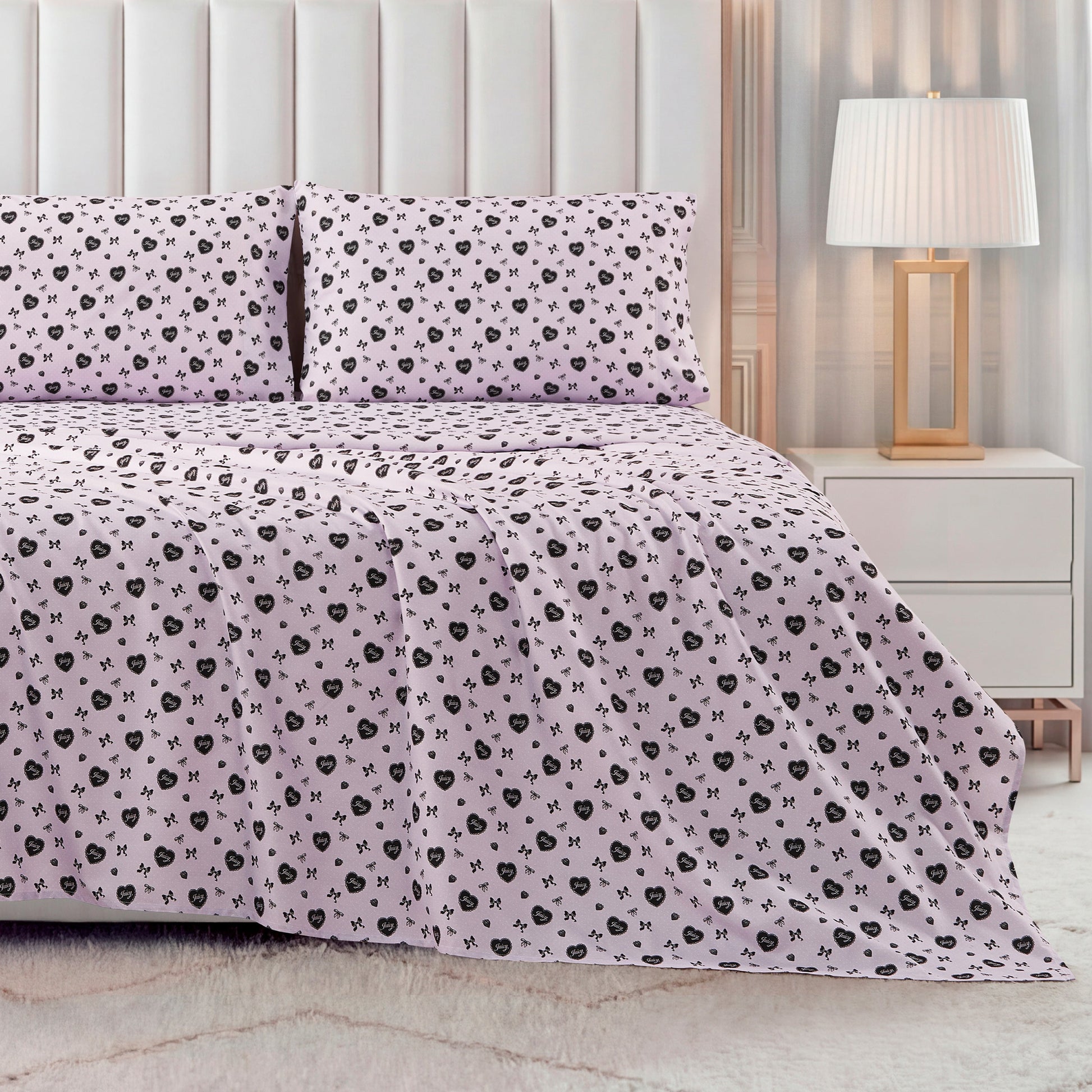 A modern bedroom featuring a neatly made bed with the Printed Microfiber Sheet Set in pale pink with a black floral pattern, matching pillows, a white upholstered headboard, plus a white nightstand and lamp with a white shade.