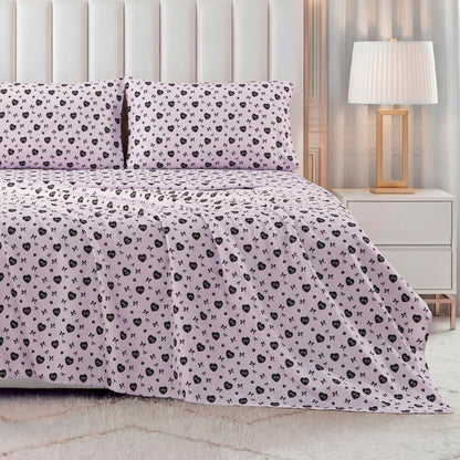 A modern bedroom featuring a neatly made bed with the Printed Microfiber Sheet Set in pale pink with a black floral pattern, matching pillows, a white upholstered headboard, plus a white nightstand and lamp with a white shade.