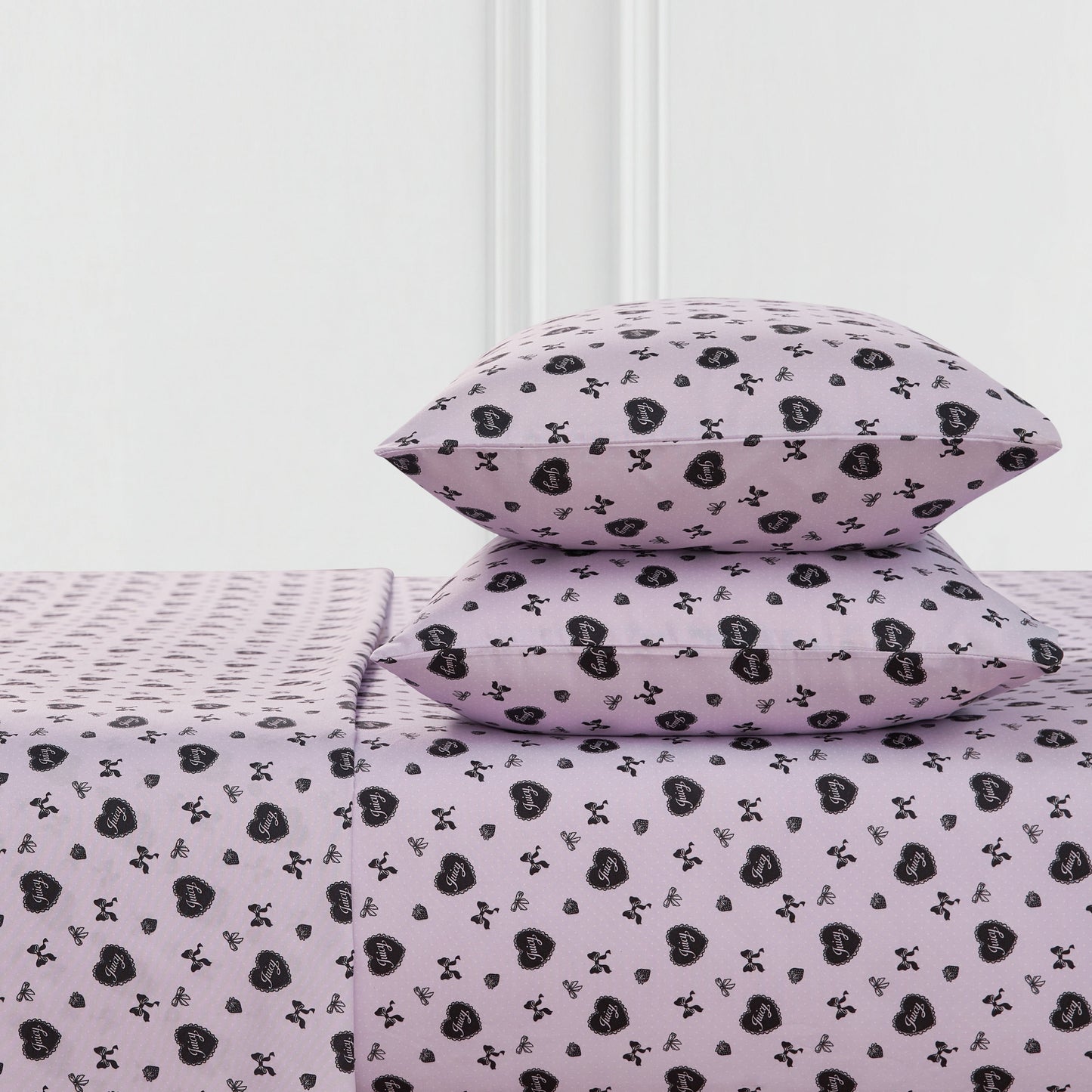 Two lavender Printed Microfiber Sheet Set pillows and a matching bedsheet, all with a repeating black heart and crossbones pattern, are neatly arranged against a white wall.