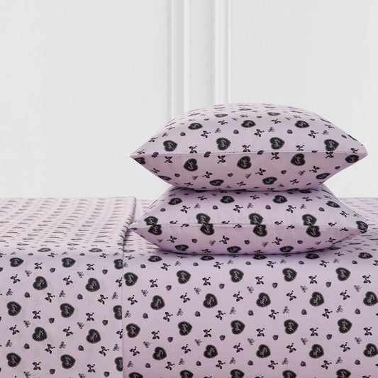 Two lavender Printed Microfiber Sheet Set pillows and a matching bedsheet, all with a repeating black heart and crossbones pattern, are neatly arranged against a white wall.