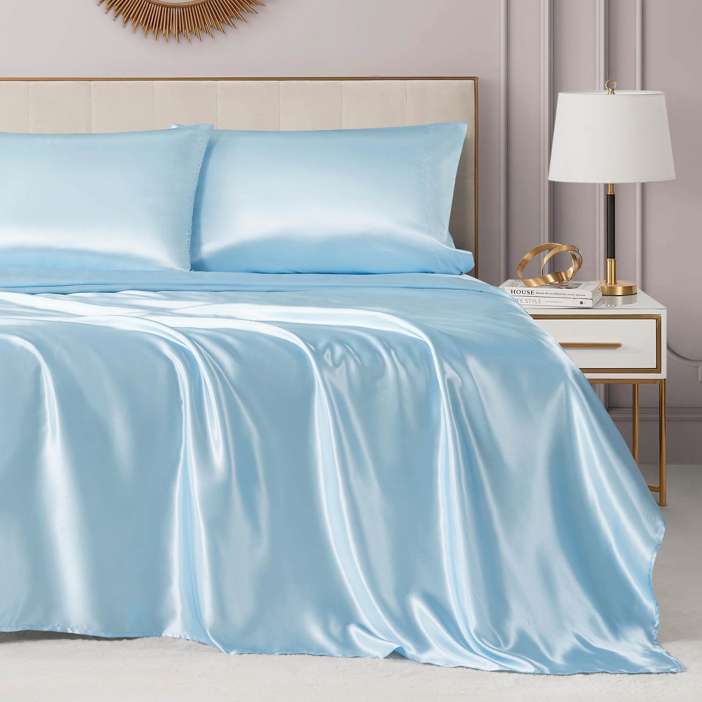 A neatly made bed features the Solid Satin Sheet Set in light blue with matching pillowcases, set against a beige upholstered headboard. Beside it is a modern white nightstand with gold accents, a white lamp, and decor items.