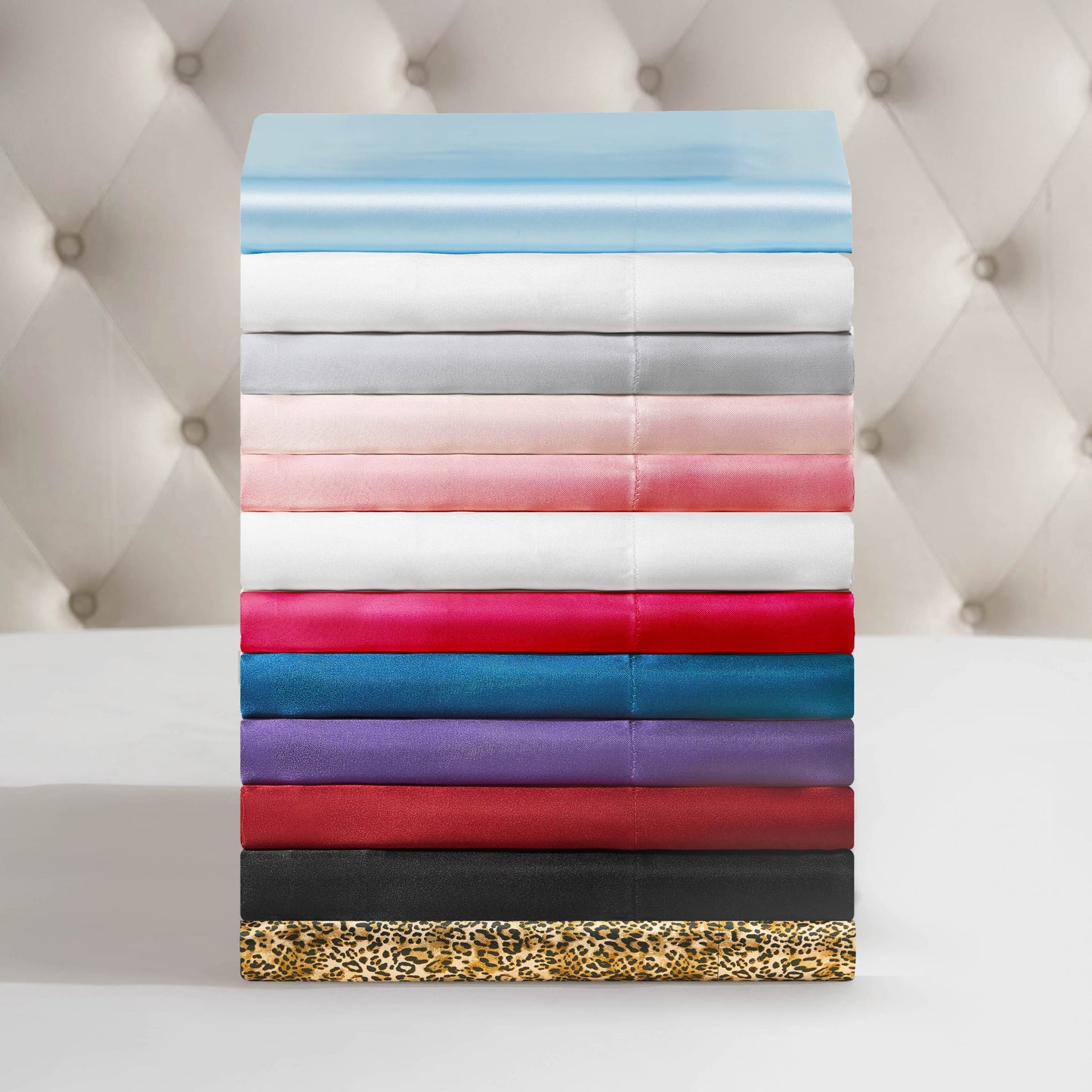 A neatly arranged stack of Solid Satin Sheet Sets in a variety of colors and patterns, including Juicy Couture-inspired pinks and leopard print, is displayed on a white surface with a tufted background.